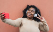© Alessandro Biascioli - Happy African woman taking selfie with mobile smartphone while listening music with wireless headphones