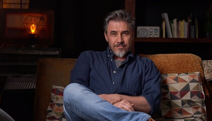 Portrait of older man at home in dark room sitting on couch. Happy smile, grey hair, bearded, . mature age, middle age, mid adult man in 50s.