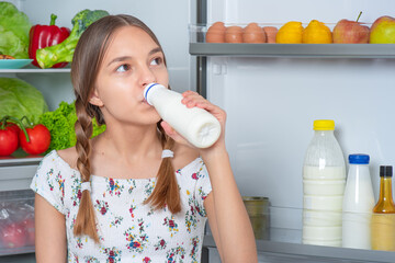 Wall Mural - Beautiful young teen girl holding bottle of milk and drinks while standing near open fridge in kitchen at home