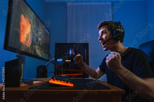 Cybersport young pro gamer happy with winning the game, feel exited, show YES hand gesture, celebrates victory in online game competition. Side view. Guy playing video game at home in his room