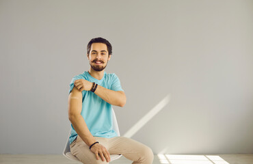 Wall Mural - Portrait of happy young man after COVID-19 shot. Cheerful guy sitting on chair shows arm after flu, coronavirus, tetanus, hepatitis, meningitis or typhoid fever vaccine. Vaccination and safety concept