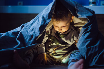 Happy smiling boy sitting in bed under the covers and playing on the smartphone in the dark. The baby's face is illuminated by a bright monitor. halloween. insomnia in children 
