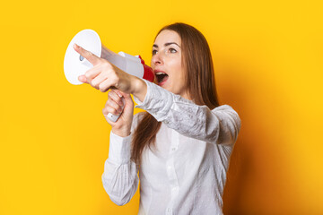 Wall Mural - Young woman in a white shirt makes an announcement in a megaphone and points a finger on a yellow background. Hiring concept, help wanted. Banner