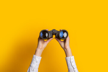 female hands hold black binoculars on a bright yellow background