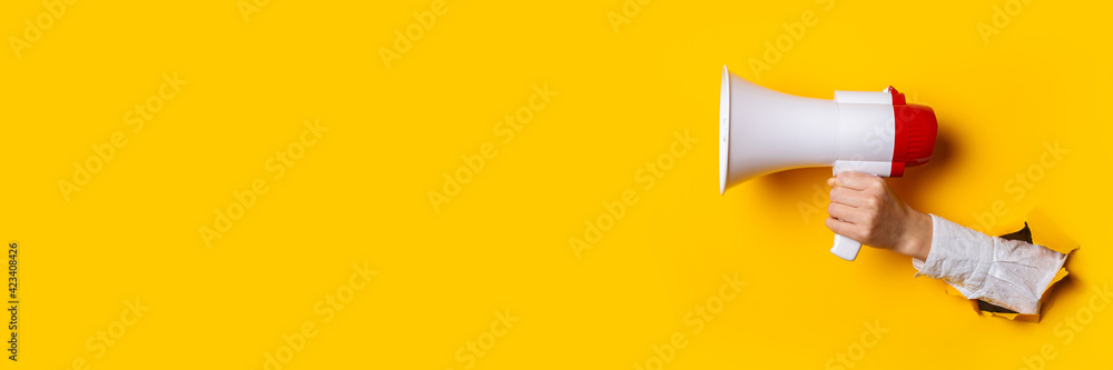 Hand holds a megaphone from a hole in the wall on a yellow background. Concept of hiring, advertising something. Banner - obrazy, fototapety, plakaty 