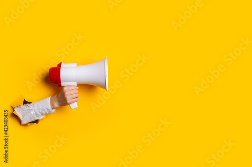 Hand holds a megaphone from a hole in the wall on a yellow background. Concept of hiring, advertising something. Banner