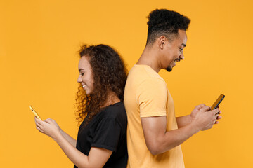 Wall Mural - Side view young couple two friends together family african students woman man 20s in black t-shirt holding using mobile cell phone stand back to back isolated on yellow background studio portrait.