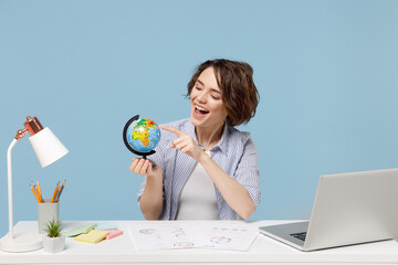 Wall Mural - Young travel agent successful secretary employee business woman in casual shirt work at white office desk with pc laptop point finger on Earth world globe isolated on pastel blue background studio.