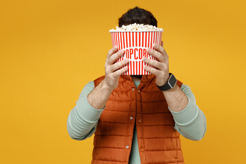 Poster - Young smiling happy cheerful fun caucasian man 20s wearing orange vest mint sweatshirt glasses holding popcorn takeaway bucket watching film comedy isolated on yellow color background studio portrait