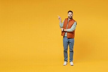 Wall Mural - Full length young smiling happy confident smiling cheerful fun caucasian man in orange vest mint sweatshirt point index finger aside on copy space area mock up isolated on yellow background studio