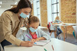 © Kostiantyn - Helpful young female teacher wearing protective face mask helping little girl doing sums. Kids studying in elementary school, sitting at the desk