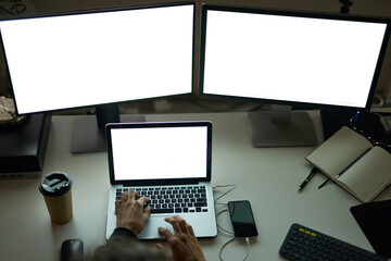 Wall Mural - Work anytime. High angle view of hands of man sitting at the table in front of many computer monitors and using laptop while working late at night