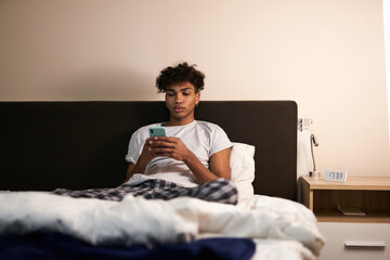Young guy with piercing chatting with friends, using smartphone in his bed at night before going to sleep