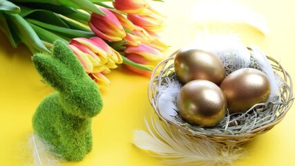 Wall Mural - Basket easter decoration: Golden eggs in basket with spring tulips, white feathers on pastel yellow background. Congratulatory easter design