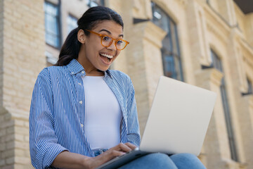 Excited African American woman using laptop computer shopping online with sales. Happy beautiful  female watching video, reading good news sitting outdoors 