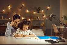 Parent Helping Child. Happy Family Doing Homework In The Evening. Mother And Daughter Working On School Assignment Sitting At Desk With Lamp In Cozy Dark Room With LED Lights. Kids Learning Concept