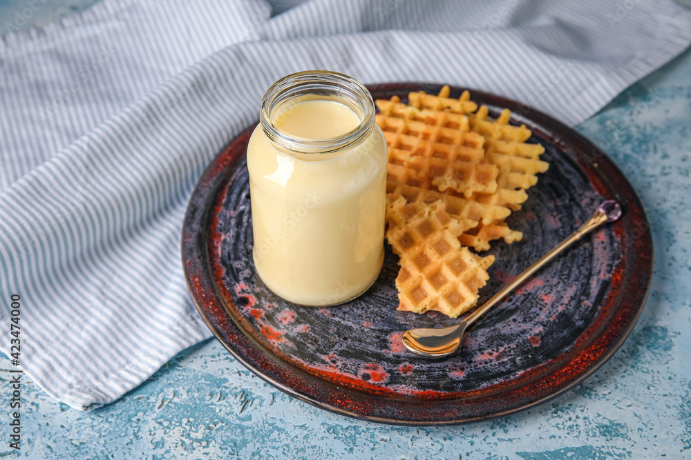 Jar with sweet condensed milk and waffles on color background