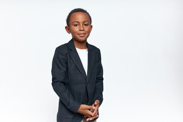 Wall Mural - Fashion, style and childhood concept. Studio image of handsome African schoolboy posing against white copy space background in formal jacket, clasping hands, looking at camera with confident smile