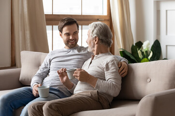 Wall Mural - Happy millennial adult Caucasian man relax on sofa in living room with senior father talk drink coffee or tea. Smiling older dad and grownup song rest at home enjoy family weekend reunion chatting.