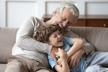 Wall Mural - Overjoyed older Caucasian grandfather relax in living room with smiling little grandson tickle and giggle. Smiling mature granddad feel playful have fun play with small 8s boy child at home together.