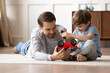 © fizkes - Loving young Caucasian father sit on floor feel playful repair fix toy car together with little 8s son. Caring happy dad play with automobiles with small preschooler boy child at home on weekend.