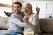 © fizkes - Overjoyed millennial Caucasian man and mature father relax at home watch football match online celebrate goal win. Happy older dad and adult grownup son enjoy team victory enjoying game on TV.