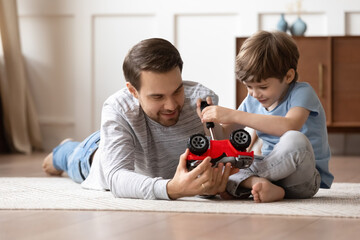 Loving young Caucasian father sit on floor feel playful repair fix toy car together with little 8s son. Caring happy dad play with automobiles with small preschooler boy child at home on weekend.