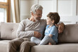 Loving old 60s Caucasian granddad sit on sofa with little curious grandson talk and chat at home. Caring mature grey-haired grandfather relax on couch with small grandchild gossip share secrets.