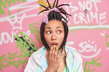 Wall Mural - Surprised teenage girl keeps hand near opened mouth feels stunned has dreadlocks looks aside with shocked expression wears stylish clothes poses against colorful painted wall. Graffiti artist