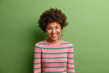 Wall Mural - Portrait of lovely young African American looks with friendly happy expression at camera expresses positive emotions being in good mood dressed casually isolated over green vivid background.