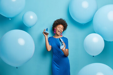 Wall Mural - Positive African American woman with curly hair keeps shoes near mouth pretends singing wears dress chooses outfit for party event isolated over blue background with inflated balloons around