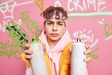 Poster - Horizontal shot of unhappy guy youngster dressed in sweatshirt holds two aerosol bottles makes creative graffiti on street wall has dissatisfied expression. Contemporary art and millenial lifestyle