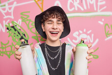 Positive Curly Hipster Guy Laughs And Has Fun With Friend While Drawing Graffiti Smiles Broadly Has Braces On Teeth Holds Aerosol Spray Bottles Wears Stylish Clothes Feels Chilly. Overjoyed Teenager