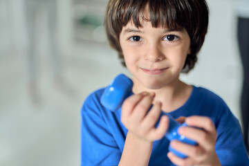 Close up portrait of adorable little latin boy smiling at camera, exercising with dumbbell in the morning at home