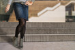 Woman wearing blue jeans skirt, black pantyhose and grey boots in city street. Fashionable spring or autumn outfit.
