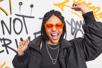 Crazy cheerful dark skinned hipster girl dances with arms raised has fun foolishes around poses against graffiti wall wears orange sunglasses black jacket belongs to youth subculture. Urban style