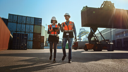 Multiethnic Female Industrial Engineer with Tablet Computer and Male Foreman Worker in Hard Hats and Orange High-Visibility Vests Walk in Container Terminal. Colleagues Talk About Logistics Business.