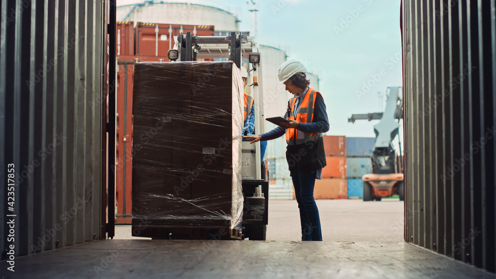 Forklift Driver Loading a Shipping Cargo Container with a Full Pallet with Boxes in Logistics Port Terminal. Latin Female Industrial Supervisor and Safety Inspector with Tablet Managing the Process. - obrazy, fototapety, plakaty 