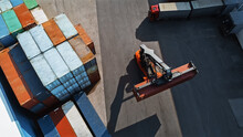 Aerial Top Down Shot Of A Container Handler Carrying A Large Red Cargo Container In A Shipyard Terminal. Driver Of The Machine Is Loading The Crate In The Logistics Center Depot.