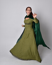 Full Length Portrait Of Red Haired Girl Wearing Celtic, Green Medieval Gown With Fantasy Velvet Cloak. Standing Pose Isolated Against A Studio Background.