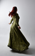 Full Length Portrait Of Red Haired Girl Wearing Celtic, Green Medieval Gown  With Shadowy Backlighting. Standing Pose Isolated Against A Studio Background.
