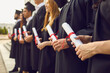 © Studio Romantic - Close up of row of students in black robes standing with traditional rolled up diplomas in hands at graduation ceremony. Happy graduates with honors in black togas celebrating their milestone event