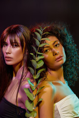 Wall Mural - Portrait of two young brunette women with professional makeup looking at camera, standing back to back, posing with green plant in neon light isolated over black background