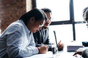 Wall Mural - Two businessmen with Asian appearance are sitting at the table signing papers and working at the computer