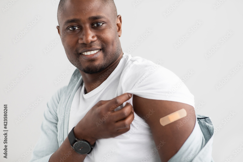 Virus protection. Happy black man holding up shirt sleeve showing the bandage after corona vaccine shot - obrazy, fototapety, plakaty 