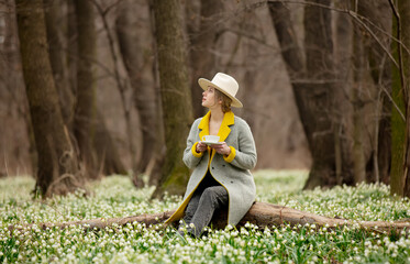 Sticker - stylish girl in gray coat with a cup in snowdrops meadow in forest