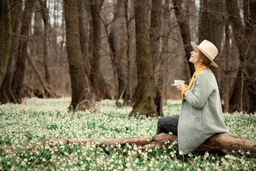 Sticker - stylish girl in gray coat with a cup in snowdrops meadow in forest