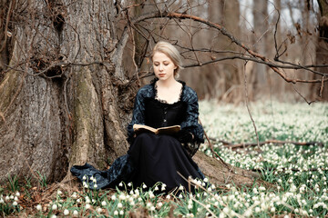 girl in vintage dress with book is sitting on meadow with snowdrops in the forest