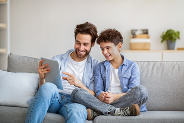 Wall Mural - Joyful father and son using tablet computer, watching videos or playing online games, spending time together at home,