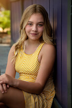 Vertical Outdoor Portrait Of A Beautiful Tween Girl With Long Blonde Hair Wearing A Yellow Dress Sitting Against A Purple Wall And Smiling At The Camera